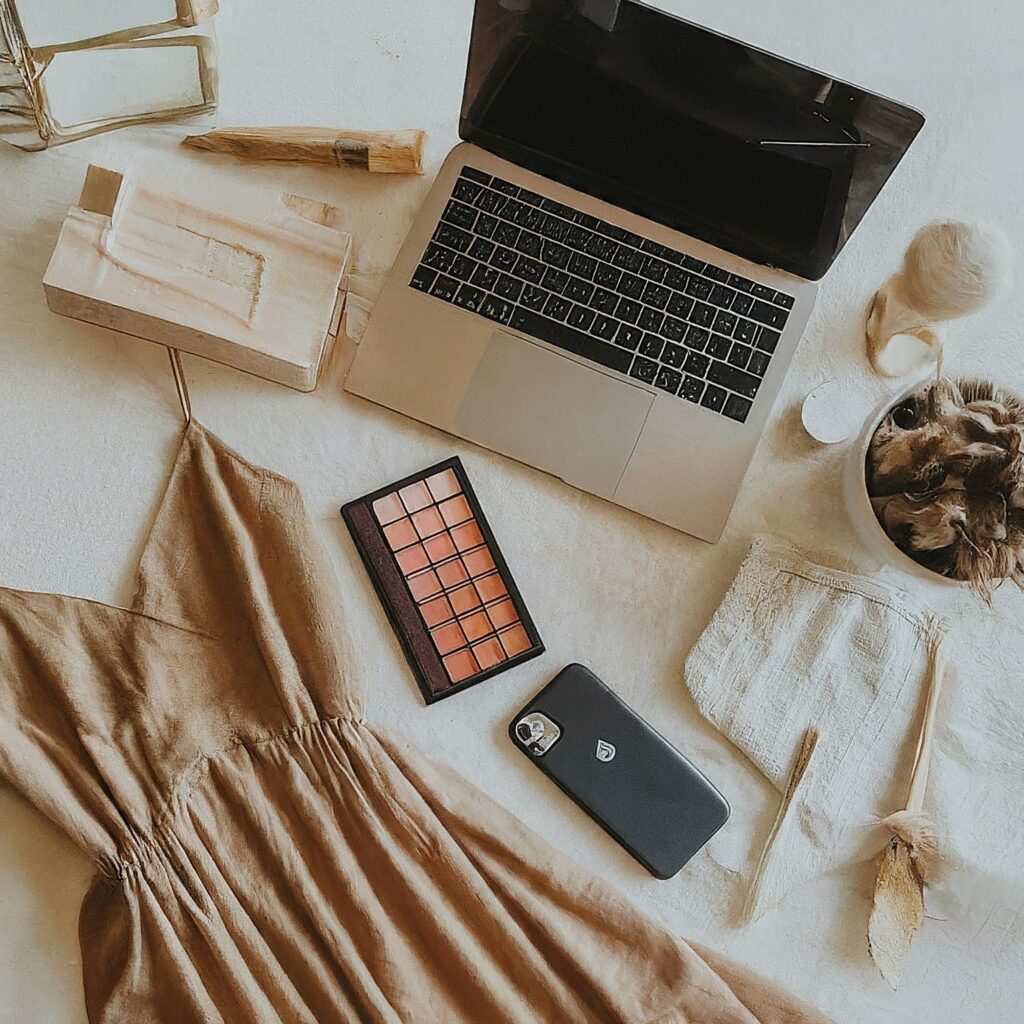 Woman shopping online on an e-commerce platform with a laptop, dress, makeup palette, and phone